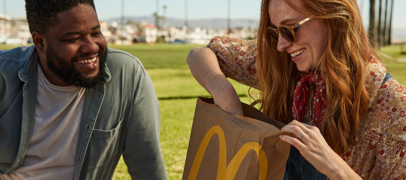 A man and woman sitting together, smiling. The woman is reaching into a McDonald's bag